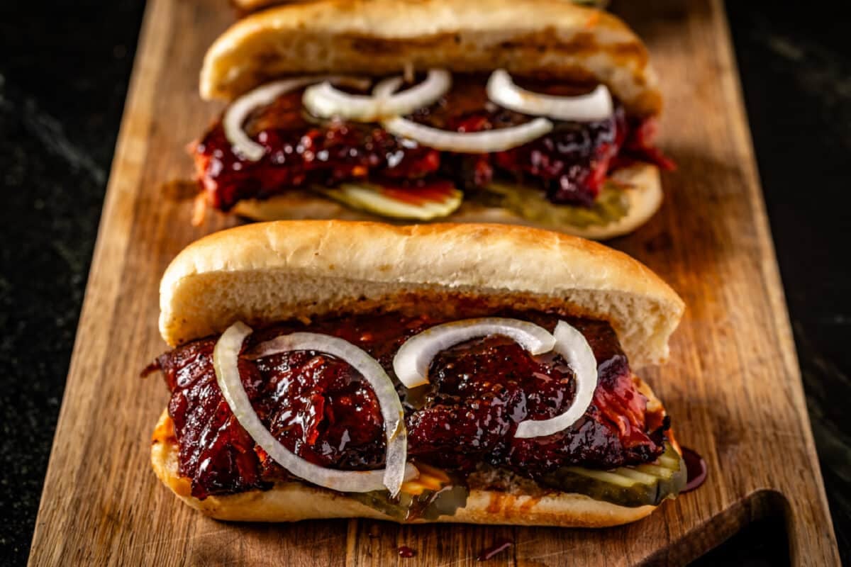 Two smoked rib sandwiches with sliced onions and pickles resting on a wooden cutting board.