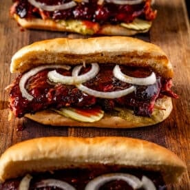 Three smoked rib sandwiches with sliced onions and pickles resting on a wooden cutting board.