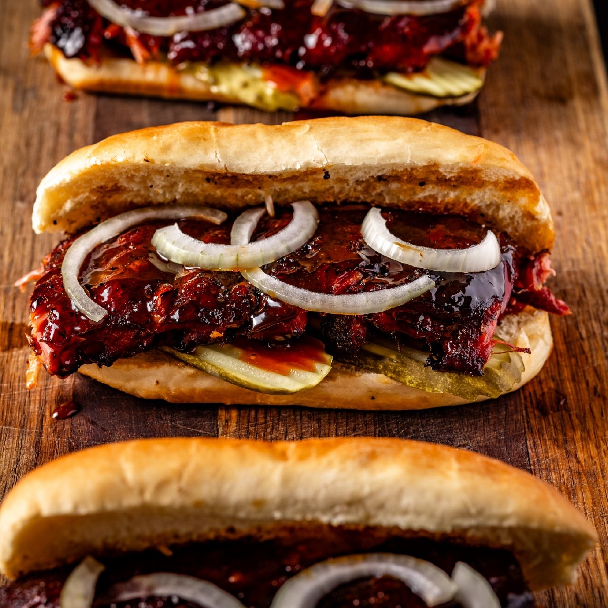 Three smoked rib sandwiches with sliced onions and pickles resting on a wooden cutting board.