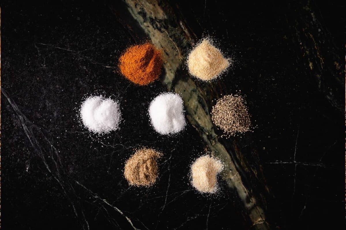 Small piles of paprika, onion powder, salt, sugar, black pepper, celery salt, and garlic powder on a marble surface.