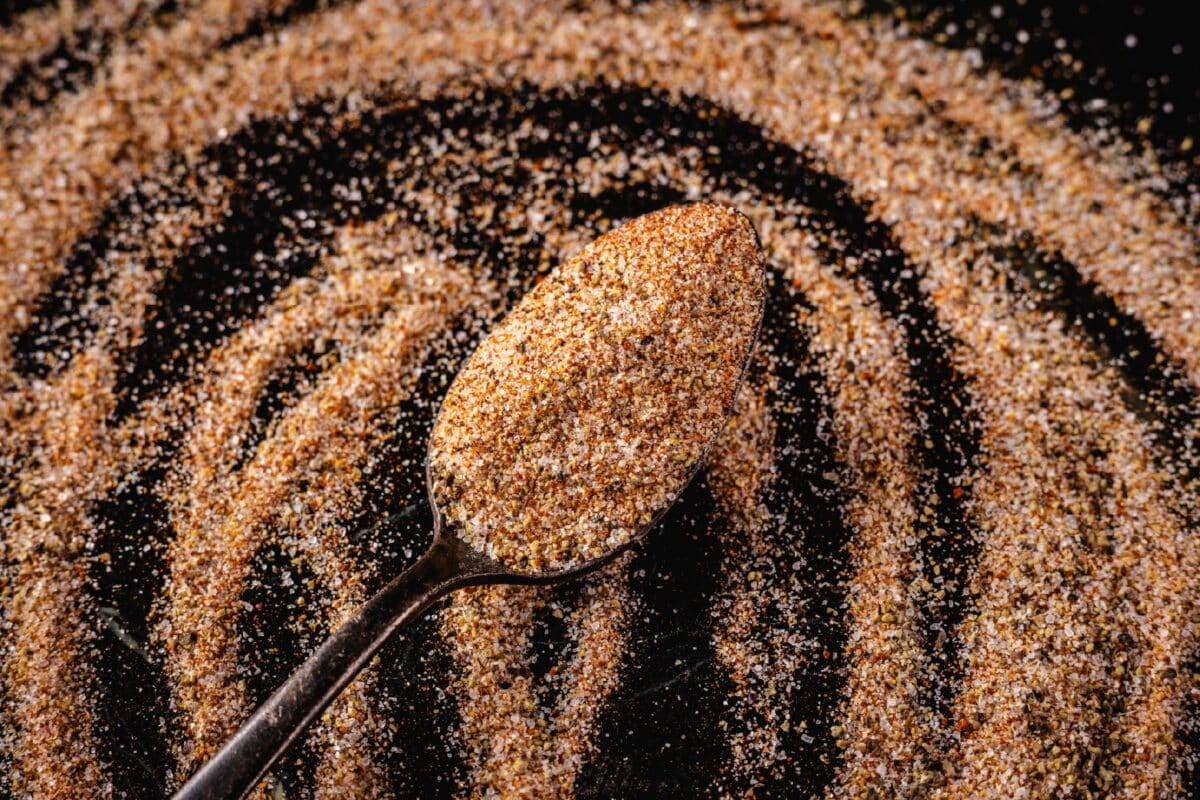 A spoon with a scoop of a seasoning blend on a dark surface dusted with the seasoning.