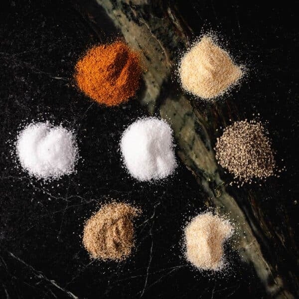 Small piles of paprika, onion powder, salt, sugar, black pepper, celery salt, and garlic powder on a dark surface.