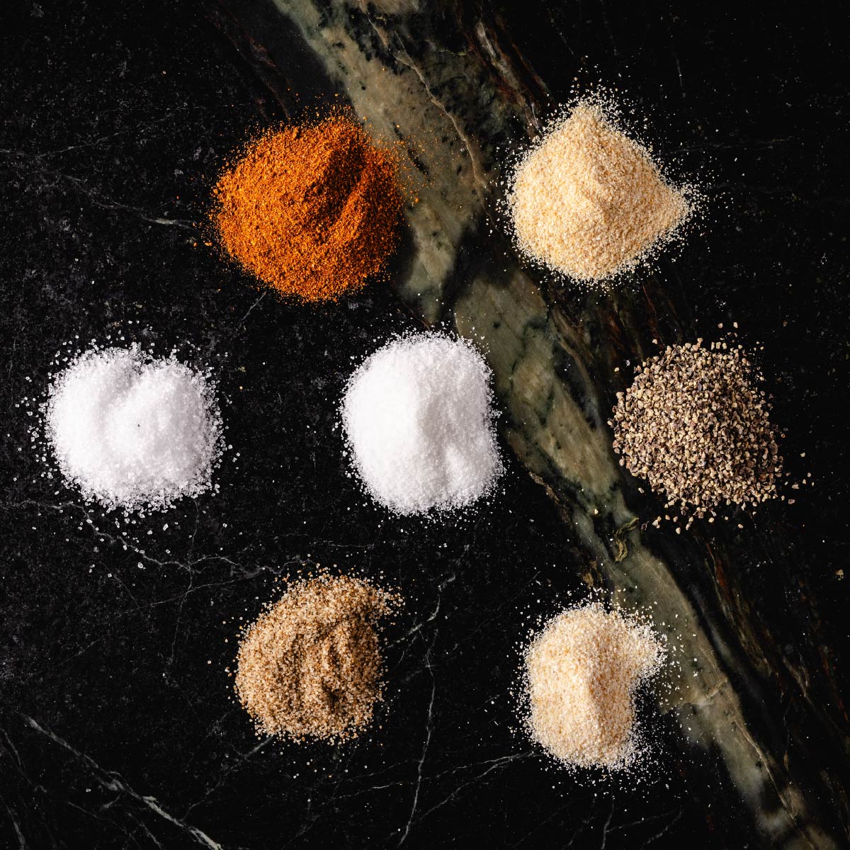 Small piles of paprika, onion powder, salt, sugar, black pepper, celery salt, and garlic powder on a dark surface.