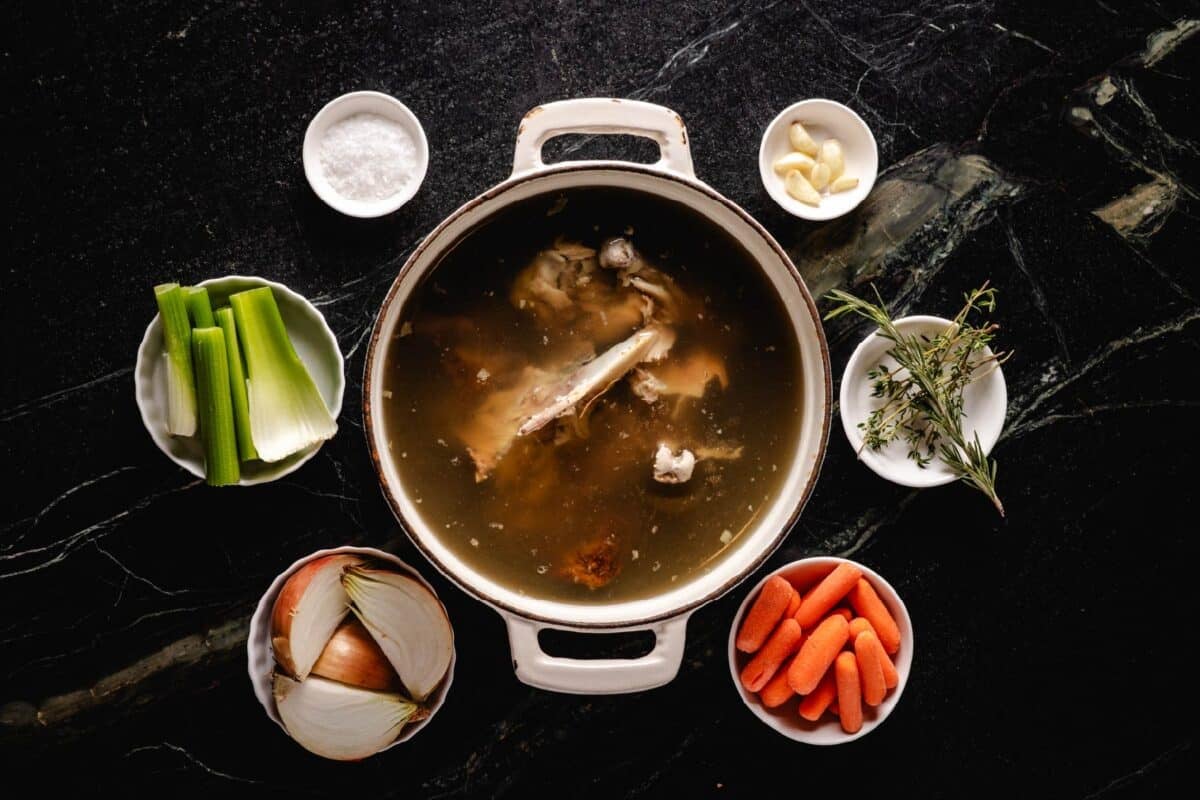 An overhead shot of a large pot of stock surrounded by small bowls of veggies and seasonings on a marble surface.