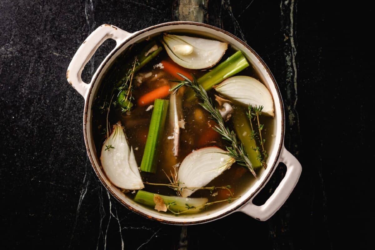 An overhead shot of a large, white pot filled with broth, onions, celery, carrots, and herbs.