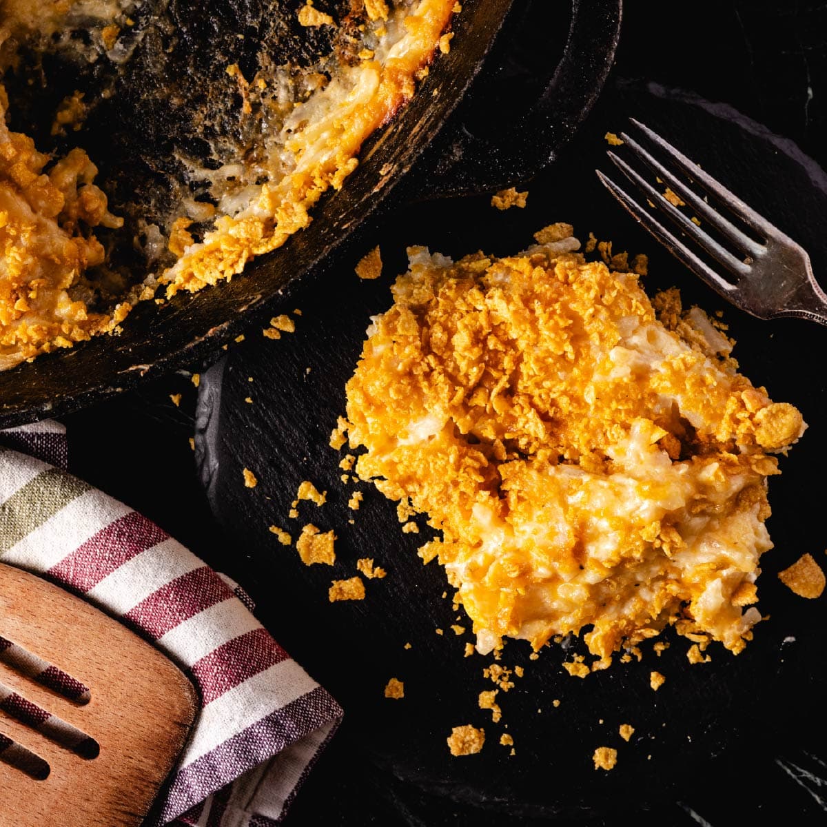 A plate with potatoes and a fork, a skillet of potatoes, and a cloth napkin with a wooden spatula resting on it.