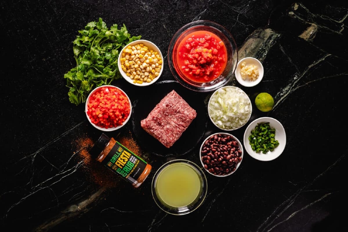 Raw ground beef on a black plate surrounded by bowls of corn, beans, tomatoes, and other ingredients.