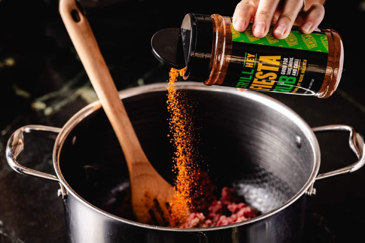A chef's and sprinkling Hey Grill Hey Fiesta Rub into a large metal pot.