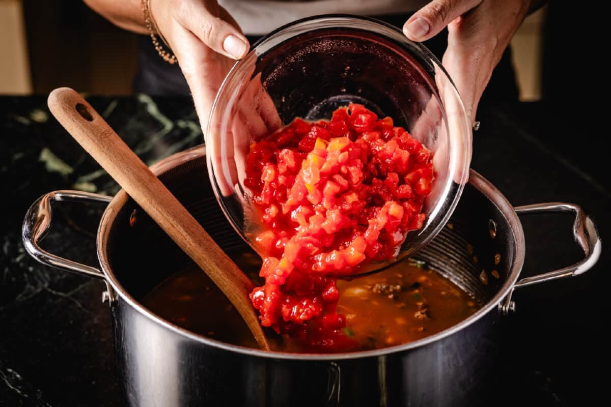 A chef's hands pouring diced tomatoes from a clear glass bowl into a large metal pot.