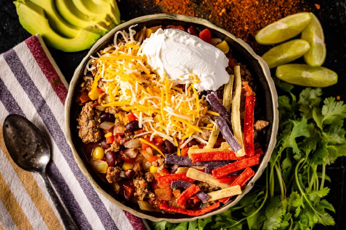 An overhead shot of a bowl of taco soup next to a cloth napkin with a spoon, and sliced avocado.
