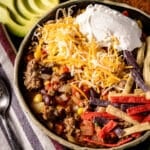 A closeup shot of a bowl of taco soup next to a cloth napkin with a spoon, and sliced avocado.