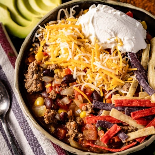 A closeup shot of a bowl of taco soup next to a cloth napkin with a spoon, and sliced avocado.