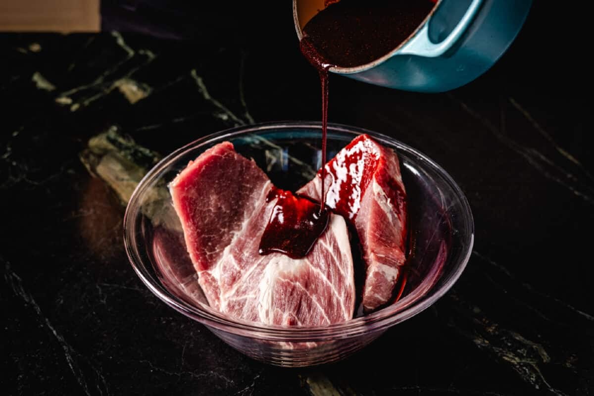 Sauce pouring from a pot into a glass bowl of raw pork.