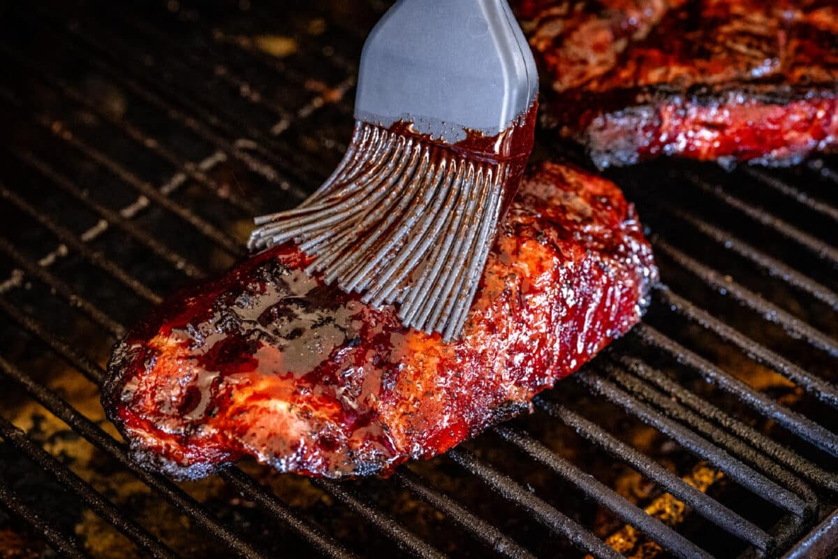 A basting brush applying sauce to pork sitting on the grates of a grill.