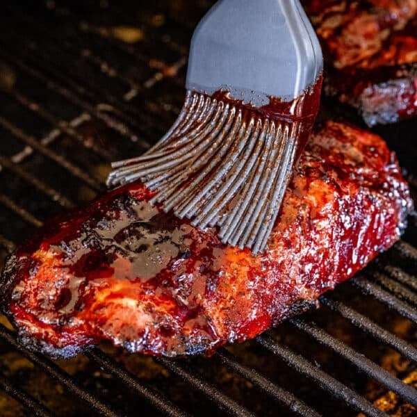 A basting brush applying char siu sauce to pork sitting on the grates of a grill.